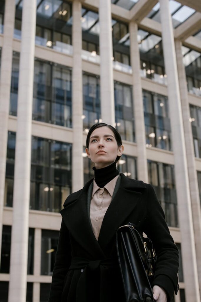 Professional woman in black coat standing in front of modern skyscraper, looking determined.